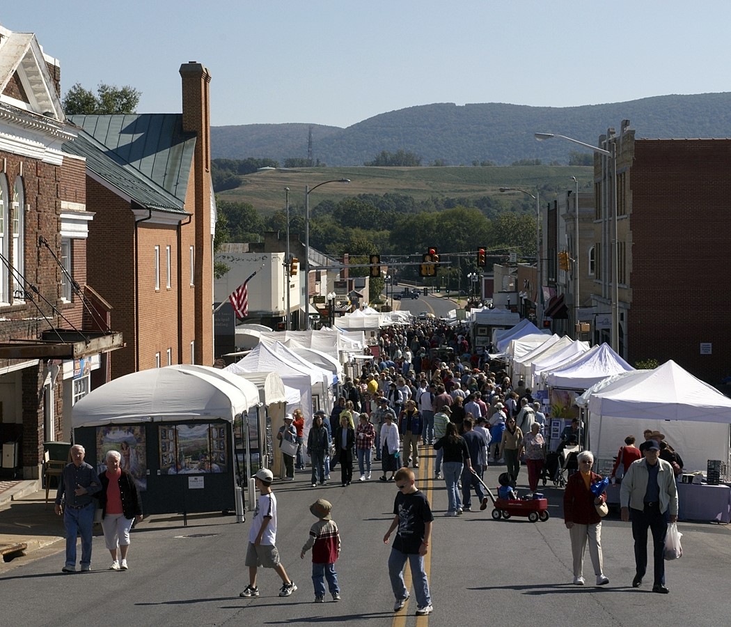 General view of a wide street in a town in the mountains, flanked by white festival tents amongst which a big crowd walk by.